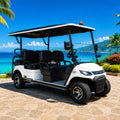 A white six-passenger golf cart with a black roof and black seats is parked on a stone patio in front of a tropical resort with palm trees, turquoise ocean, and a covered pavilion.