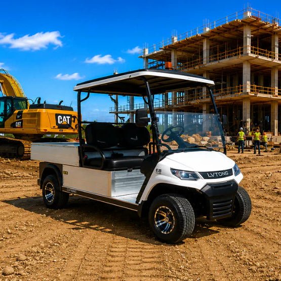 A white two-seat utility cart with a black roof is parked on a dirt construction site in front of a building under construction, with a yellow CAT excavator and workers visible in the background under a blue sky.